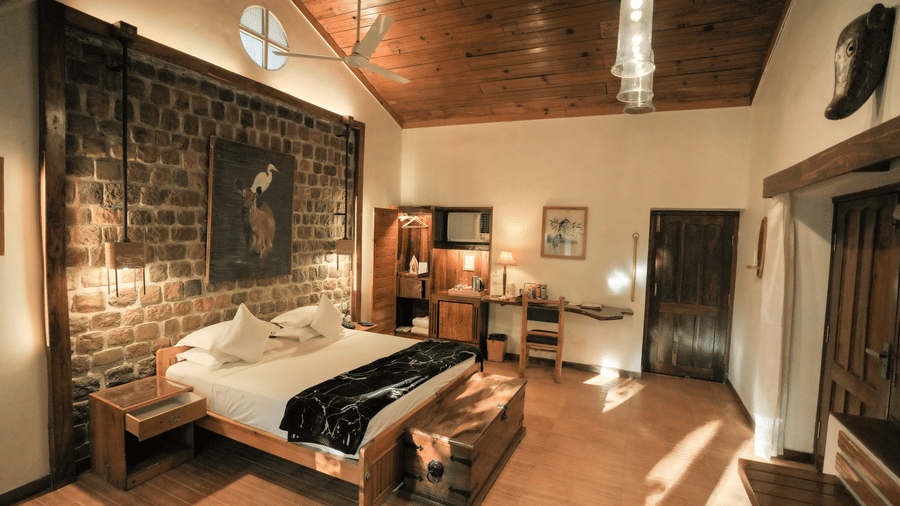 View of a room at The Den Corbett Resort and Spa Kumeria with a king bed, rustic stone wall, wooden ceiling, pendant lights, and warm ambient lighting.