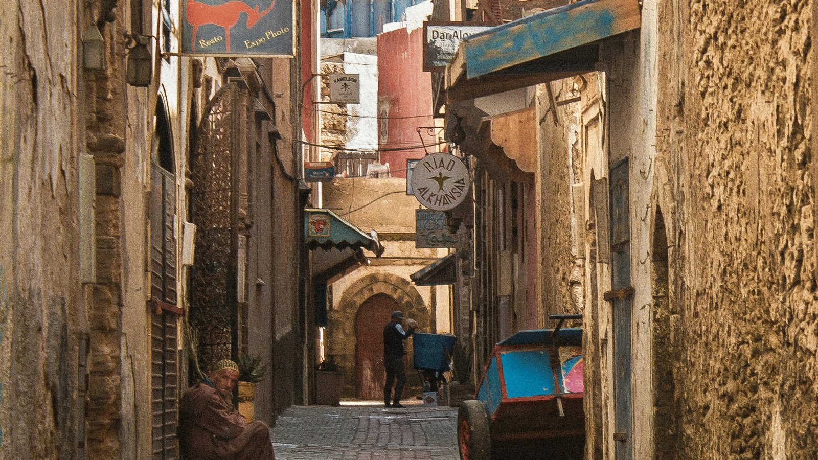 A narrow cobblestone street lined with old buildings, local shops, and a person walking with a cart.
