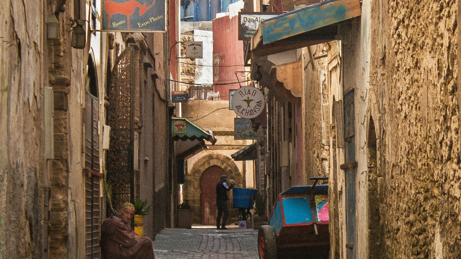 A narrow cobblestone street lined with old buildings, local shops, and a person walking with a cart.