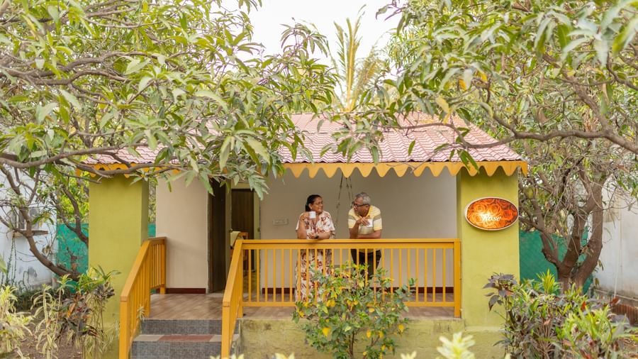 An outdoor view of a small yellow and green cottage with a veranda and 2 people standing outside, at Daksh Resort & Amusement Park, Sasan Gir.
