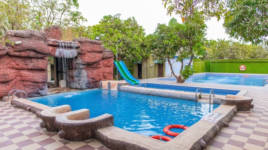 A view of the swimming pool with an artificial waterfall cascading down the rock surrounded by greenery at Daksh Resort & Amusement Park, Sasan Gir.