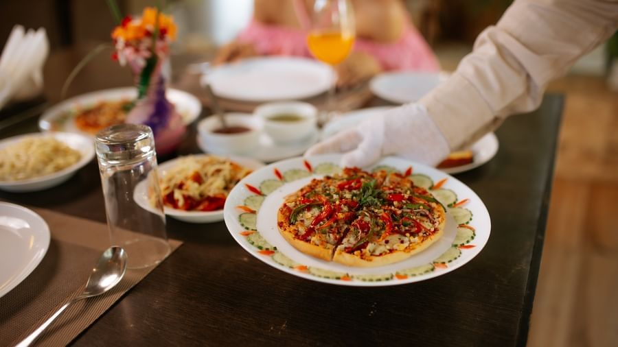 A close-up shows a waiter's gloved hands serving a plate of food onto a black dining table, at Daksh Resort & Amusement Park, Sasan Gir.