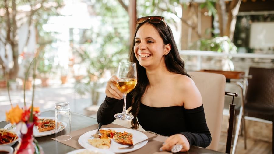 A smiling woman in a black top sits at an outdoor table with food, at Daksh Resort & Amusement Park, Sasan Gir.