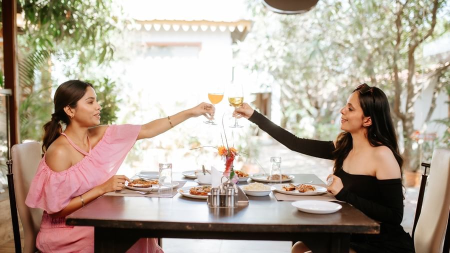 2 women are clinking wine glasses in a toast while seated at an outdoor dining table, at Daksh Resort & Amusement Park, Sasan Gir.