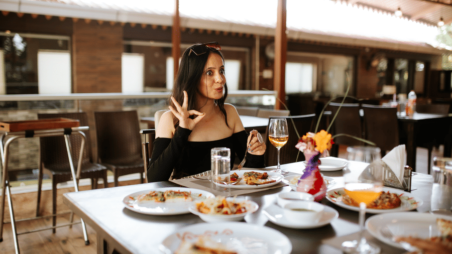 A woman in black sits at an outdoor table with food and a drink in a wine glass, at Daksh Resort & Amusement Park, Sasan Gir.