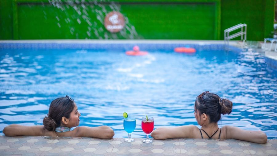 2 people relaxing at the edge of a swimming pool with drinks at Daksh Resort & Amusement Park, Sasan Gir.