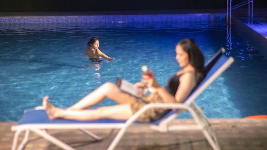A woman lies on a lounge chair by a dark pool at night, holding a drink at Daksh Resort & Amusement Park, Sasan Gir.