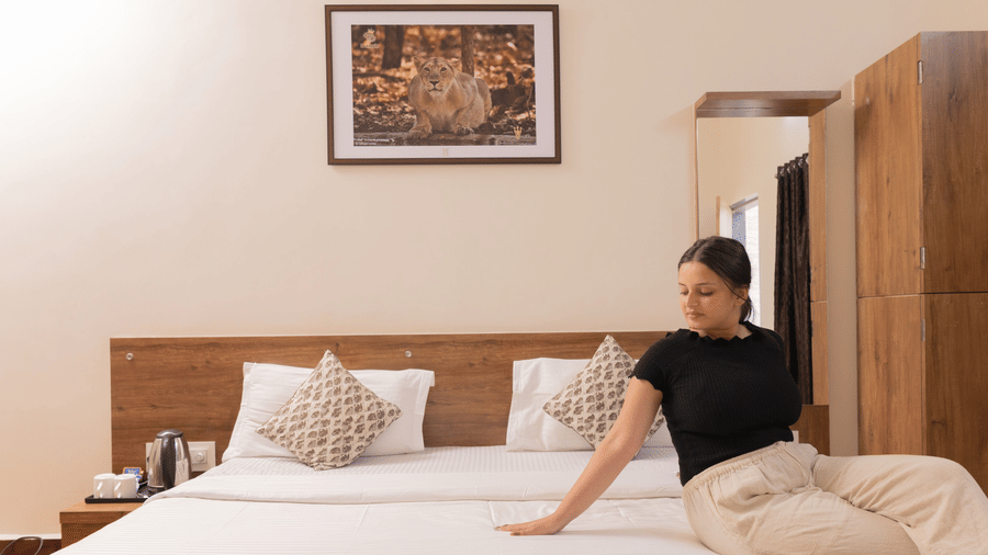 A woman sitting on a made bed, facing sideways in a guest room featuring a wardrobe, bedside table and a wall art at Daksh Resort & Amusement Park, Sasan Gir.