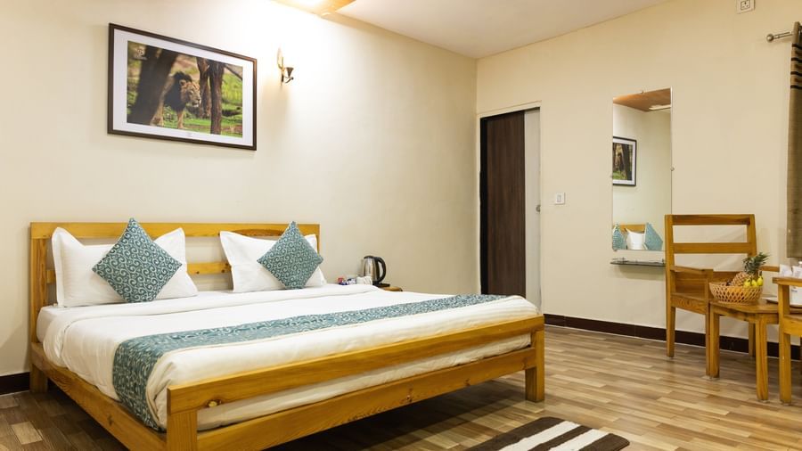 A clean room with a large bed, blue accents, a mirror, a wall art and a small chair in the corner, at Daksh Resort & Amusement Park, Sasan Gir.
