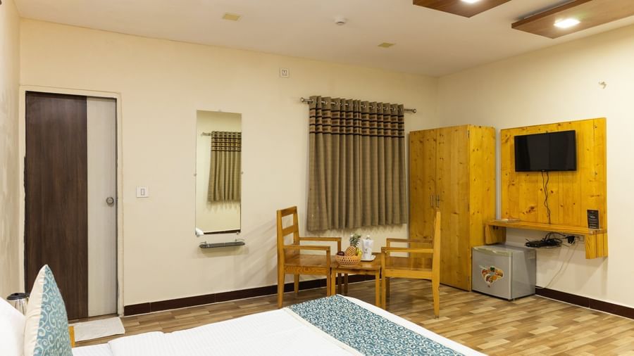 A wide view of a spacious room featuring a king size bed, a wardrobe, a wooden table with chairs, and a TV, at Daksh Resort & Amusement Park, Sasan Gir.