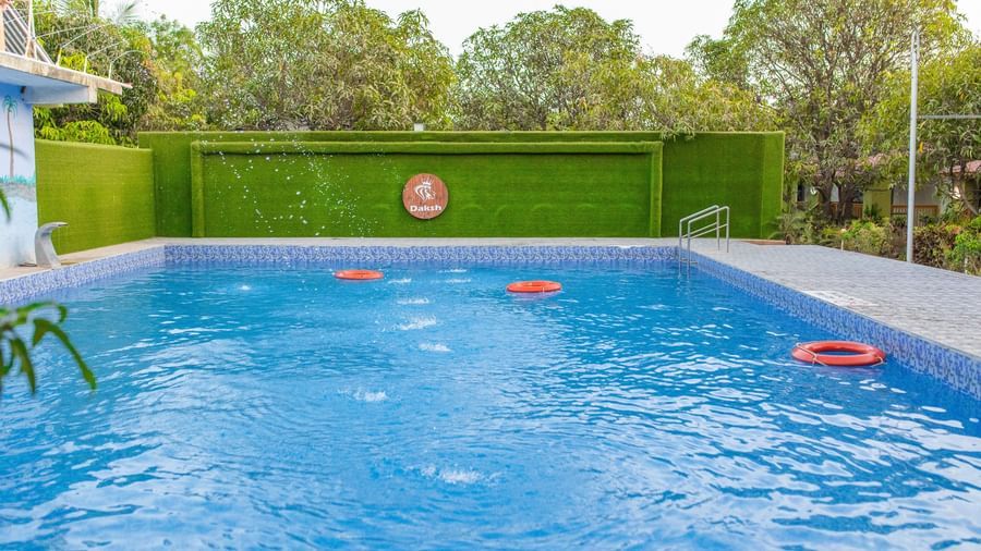 A large blue swimming pool with floating rings, bordered by trees and a green wall, at Daksh Resort and Amusement Park.