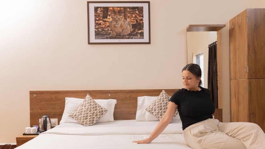 A woman sitting on a made bed, facing sideways in a guest room featuring a wardrobe, bedside table and a wall art at Daksh Resort & Amusement Park.