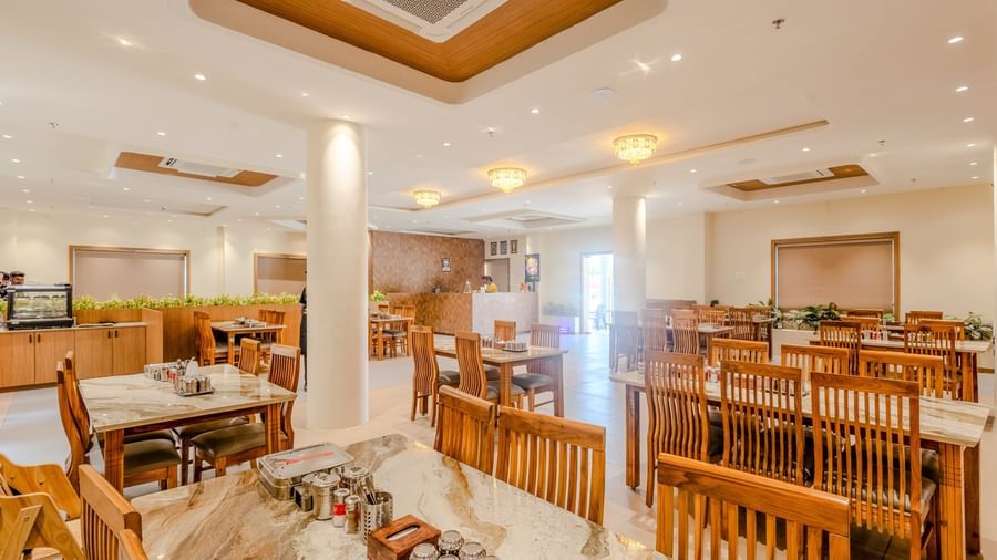 A wide-angle shot of the restaurant at Daksh The Grand Goverdhan, Dwarka, with well-arranged dining tables along with the entrance door to the place at the far end of the room.