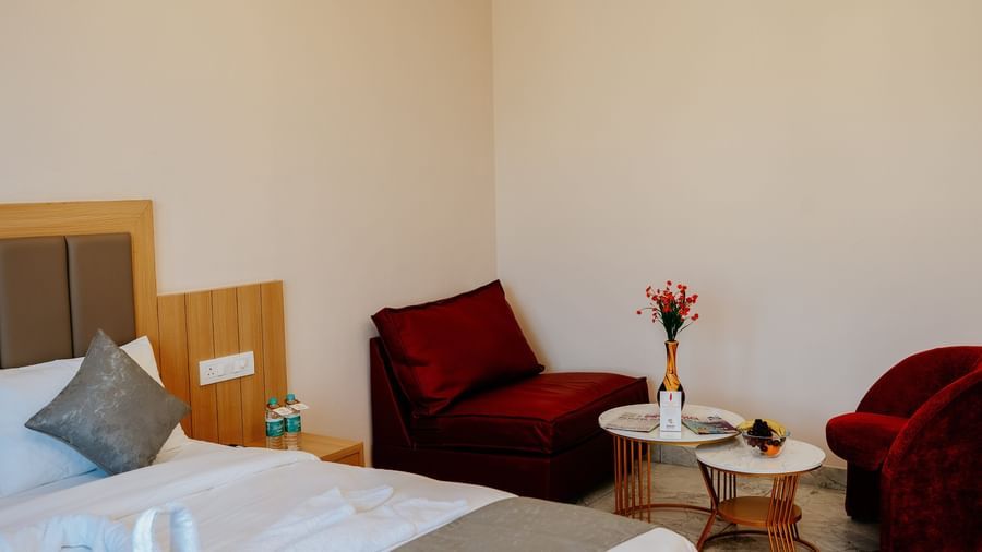 Side-angle view of the coffee corner of the Bliss room featuring 2 red-coloured arm chairs along with a decorated coffee table at Daksh The Grand Goverdhan, Dwarka.