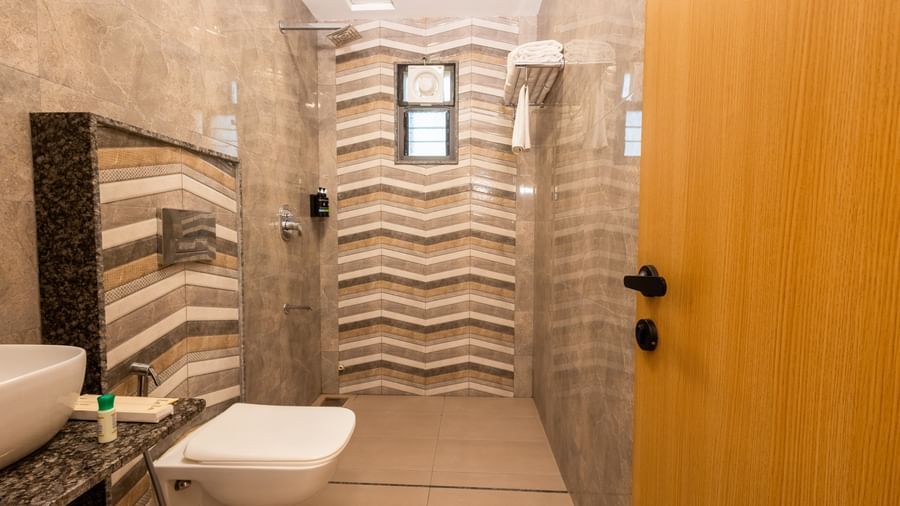 A modern bathroom at Daksh The Grand Gowardhan, Dwarka, showcasing its toilet, patterned wall tiles, and a wooden door.