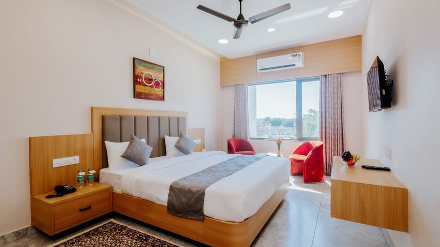 A spacious bedroom at Daksh The Grand Gowardhan, Dwarka, featuring a comfortable double bed opposite to a wall mounted television, and a common seating area at the far end of the room.