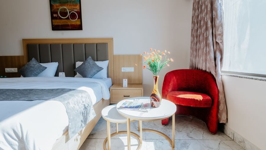 A brightly lit corner of a bedroom at Daksh The Grand Gowardhan, Dwarka, featuring a bed, a red armchair, and a small coffee table.