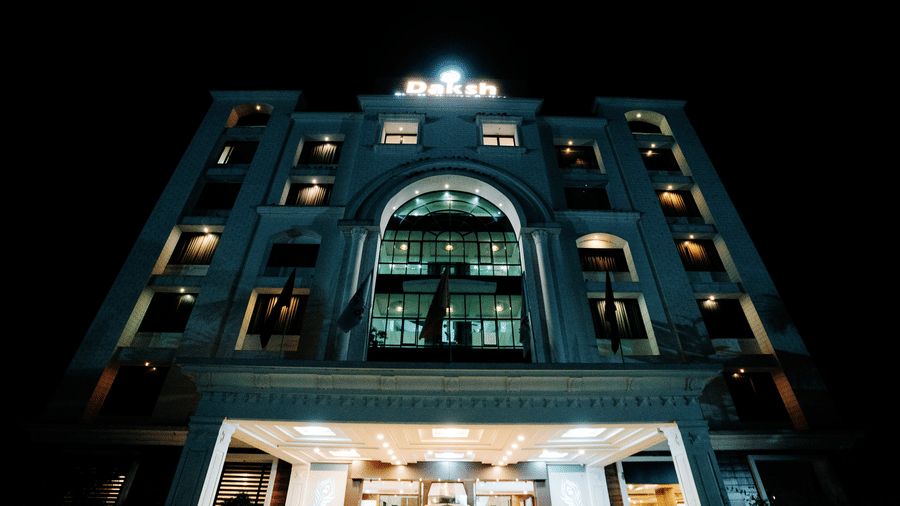 Dramatic low-angle night-time shot of the hotel's brightly lit white exterior. The arched central window and entrance are prominently illuminated at Daksh The Madhuvan Suites, Dwarka.