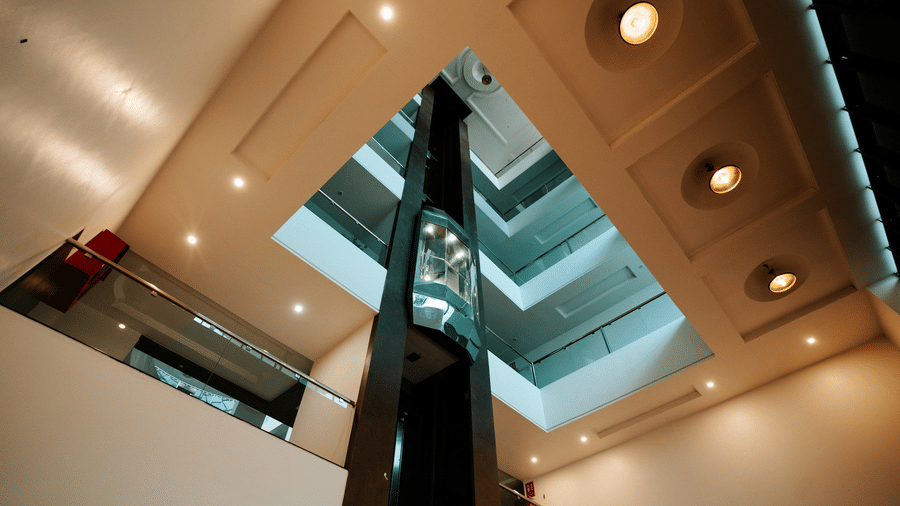A striking architectural interior shot looking up. Features a multi-layered ceiling with embedded lighting and reflective mirrored surfaces, possibly around an elevator at Daksh The Madhuvan Suites, Dwarka.