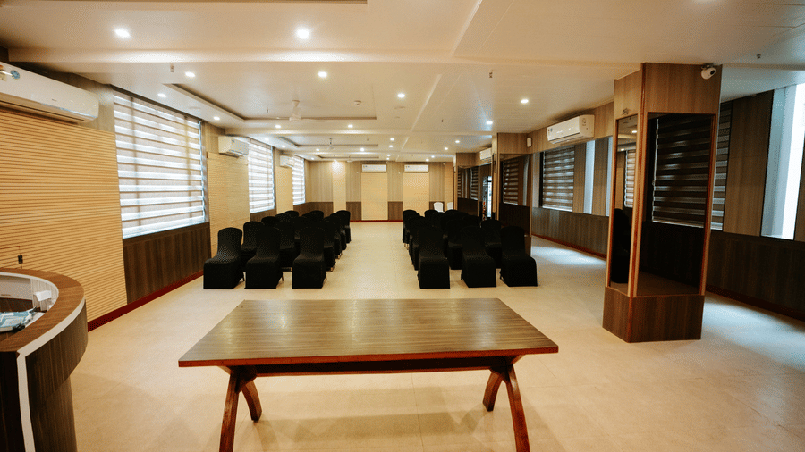 View of the long hall showing a large, dark wood meeting table in the foreground and rows of black chairs further back at Daksh The Madhuvan Suites, Dwarka.