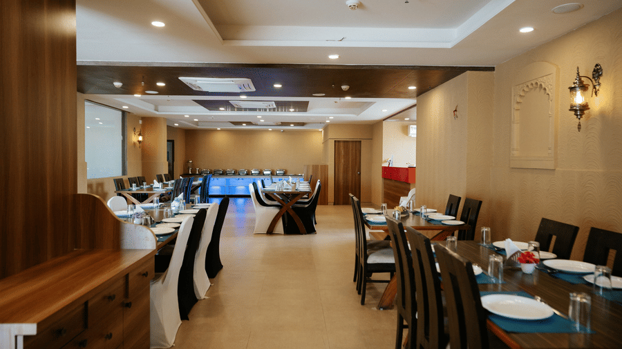 Perspective view of the long dining hall, showing multiple set tables and a glimpse of the illuminated buffet counter at Daksh The Madhuvan Suites, Dwarka.