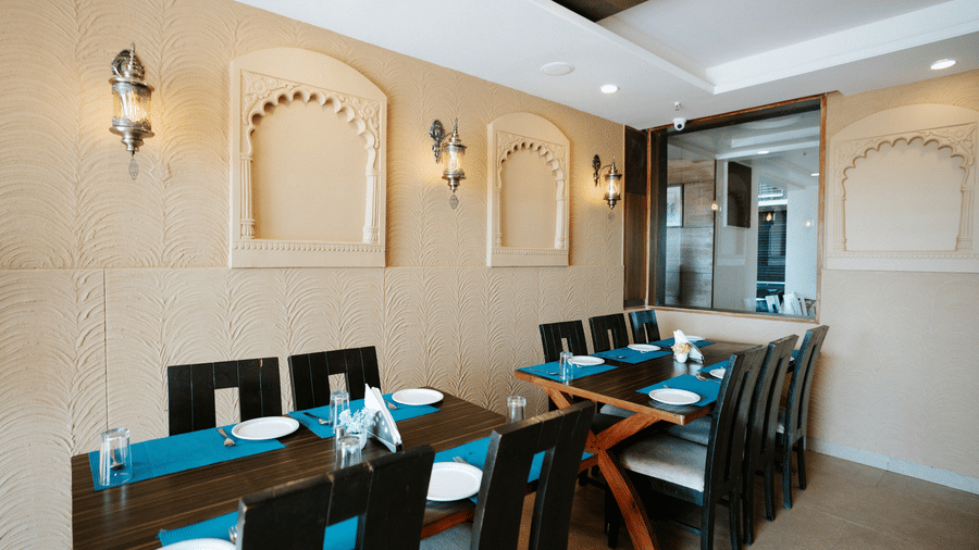 Restaurant area with tables set with blue runners, decorative beige wall arches, and traditional style wall lighting at Daksh The Madhuvan Suites, Dwarka.