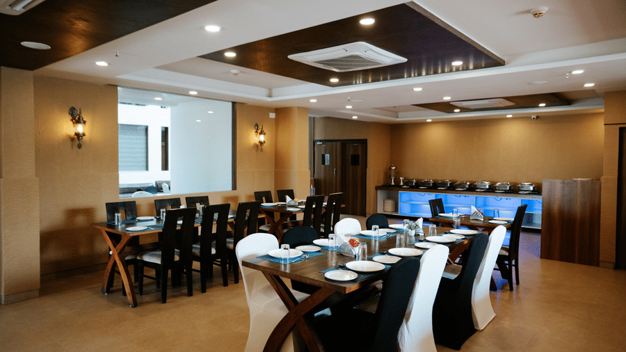 Wide shot of a well-set dining room with dark wood tables, white and black chairs, and beige patterned walls at Daksh The Madhuvan Suites, Dwarka.