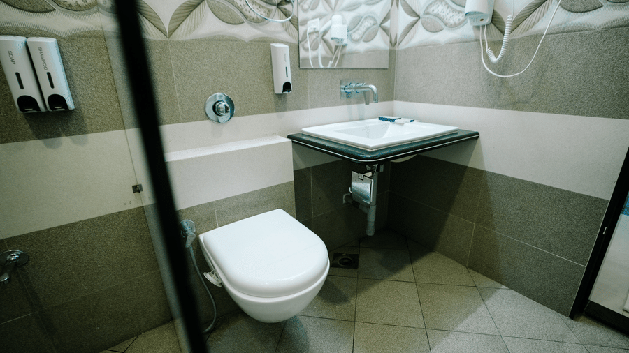 View of the clean, modern bathroom showing the square wall-mounted basin and the toilet, framed by light-coloured patterned tiles at Daksh The Madhuvan Suites, Dwarka.