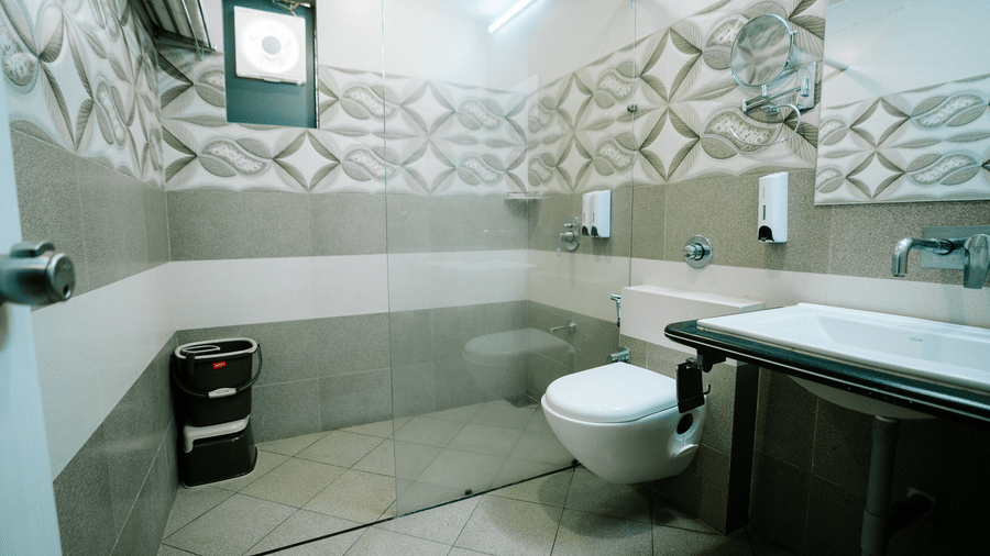 Modern bathroom with light tiled floors, a toilet, and dark vanity unit. The walls feature a patterned relief tile design at Daksh The Madhuvan Suites, Dwarka.