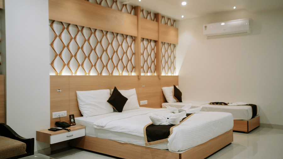 Close-up view of the bed and bedside table. White linen, dark pillows, and a wooden headboard with geometric lattice detail at Daksh The Madhuvan Suites, Dwarka.