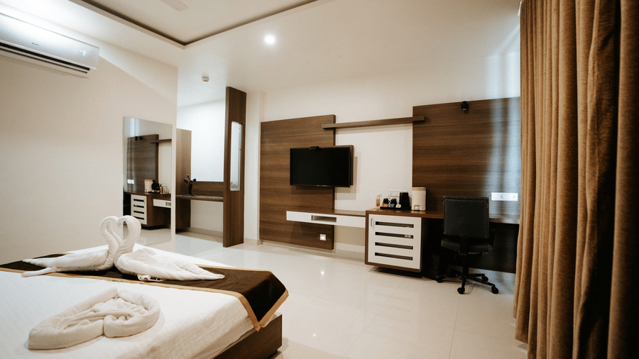 Panoramic view of the upscale hotel room, showing the bed, dark wood furniture, media console, and rich brown curtains at Daksh The Madhuvan Suites, Dwarka.