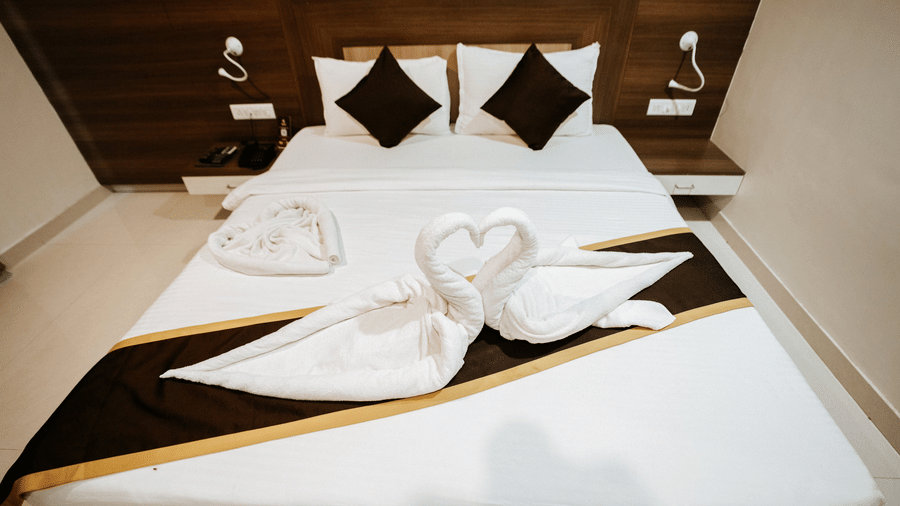 Modern, well-lit hotel room with a large bed, brown and gold accents, TV, desk, AC unit, and elegant, floor-length curtains at Daksh The Madhuvan Suites, Dwarka.