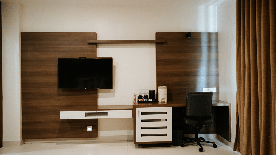 Double hotel bed with white linen, a brown and gold runner, decorative pillows, and towel swans. Wooden headboard at Daksh The Madhuvan Suites, Dwarka.