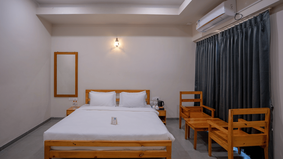 A clean and airy guest room with a double bed, wooden seating area, and dark curtains framing a large window - Daksh The Nirvana Retreat, Pavagadh