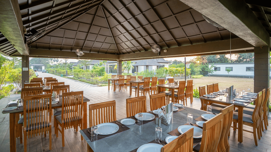 An open-air dining area with several wooden tables neatly set with white plates under a high vaulted ceiling - Daksh The Nirvana Retreat, Pavagadh