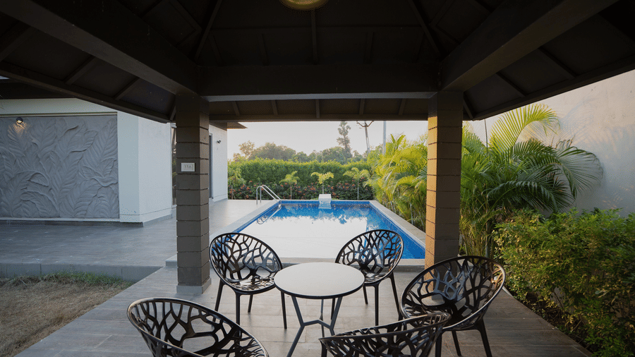 A shaded poolside patio area with a glass-top table and four black stylish chairs overlooking the water - Daksh The Nirvana Retreat, Pavagadh