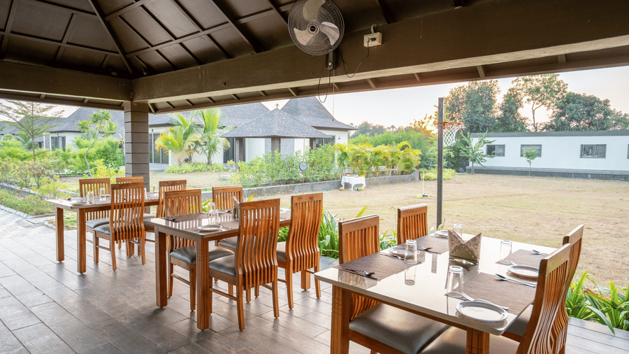 A close-up view of neatly arranged wooden dining tables in an airy, covered outdoor restaurant area - Daksh The Nirvana Retreat, Pavagadh
