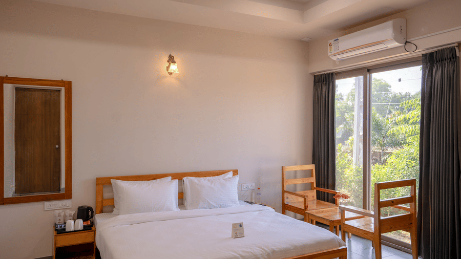 A cosy bedroom view highlighting the wooden bed frame, bedside table, and air conditioning unit under warm wall lighting - Daksh The Nirvana Retreat, Pavagadh