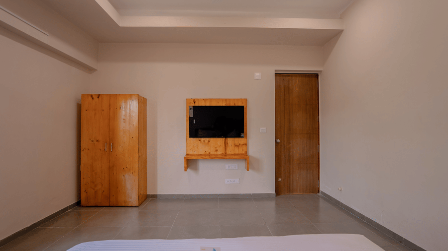 A spacious, minimalist room with smooth grey flooring, a wooden wardrobe, and a wall-mounted television - Daksh The Nirvana Retreat, Pavagadh