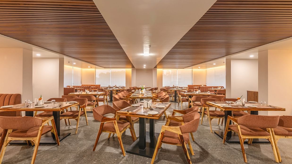 A spacious restaurant of The Bridge with wooden ceiling panels, modern lighting, and neatly arranged tables with brown chairs and elegant table settings at Clarks Inn Suites Jaipur