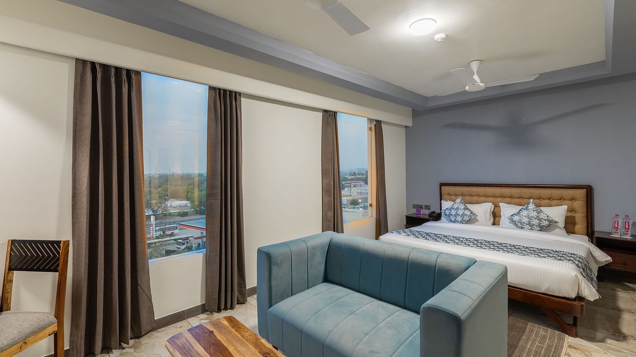 Clarks Inn Suites Jaipur, Ajmer Road featuring room with large windows, king size bed, blue sofa, wooden furniture, ceiling fans, and city view outside the curtains.