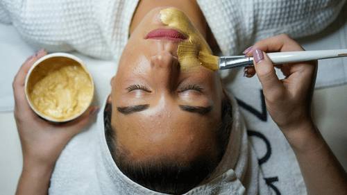 A young woman lies with her eyes closed as a beautician applies a golden-yellow face mask with a brush.
