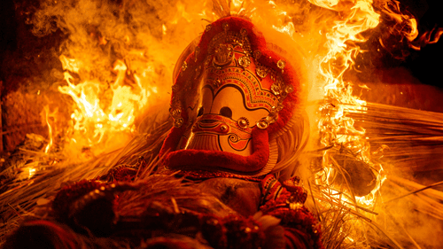 A large effigy with a traditional mask and red cloth, burning in bright orange flames at night.