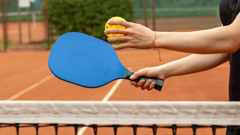 Player holding a blue pickleball paddle and tossing a yellow ball above a net, preparing to serve