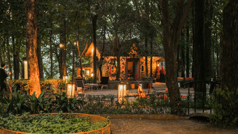 A garden with circular flower beds and warm lights at dusk, surrounded by tall trees.