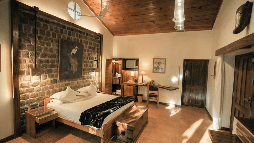 View of a room at The Den Corbett Resort and Spa Kumeria with a king bed, rustic stone wall, wooden ceiling, pendant lights, and warm ambient lighting.