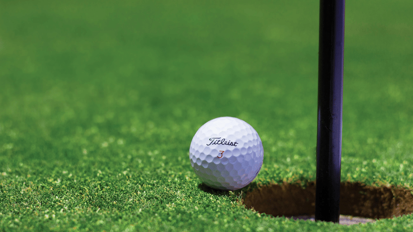 A golf ball resting next to the hole and the flag pole on a very green, well-maintained putting green.