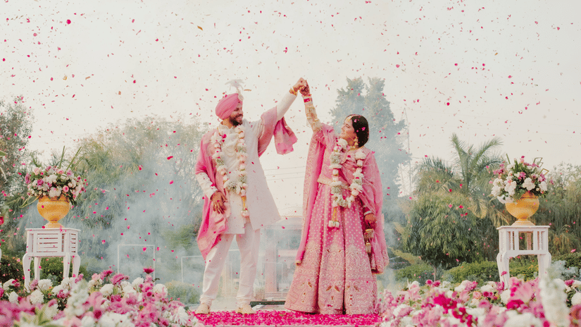 A newlywed couple holds hands and raises their arms on a stage decorated with white and pink flowers during an outdoor wedding ceremony at Umaid Palace.