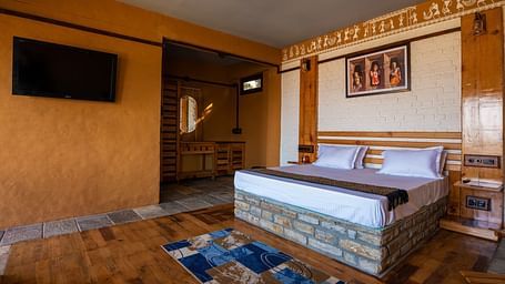 Rustic hotel room with wooden bed, wall art, TV, and patterned rug on hardwood floor. - Blusalzz Homes The Himalayan Bungalow, Almora
