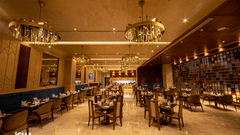 An elegant, warm-toned restaurant at The Gaurs Sarovar Premiere. Tables and chairs are set for dining. Decorative chandeliers illuminate the space. The room has a polished floor and patterned walls, creating a sophisticated atmosphere.